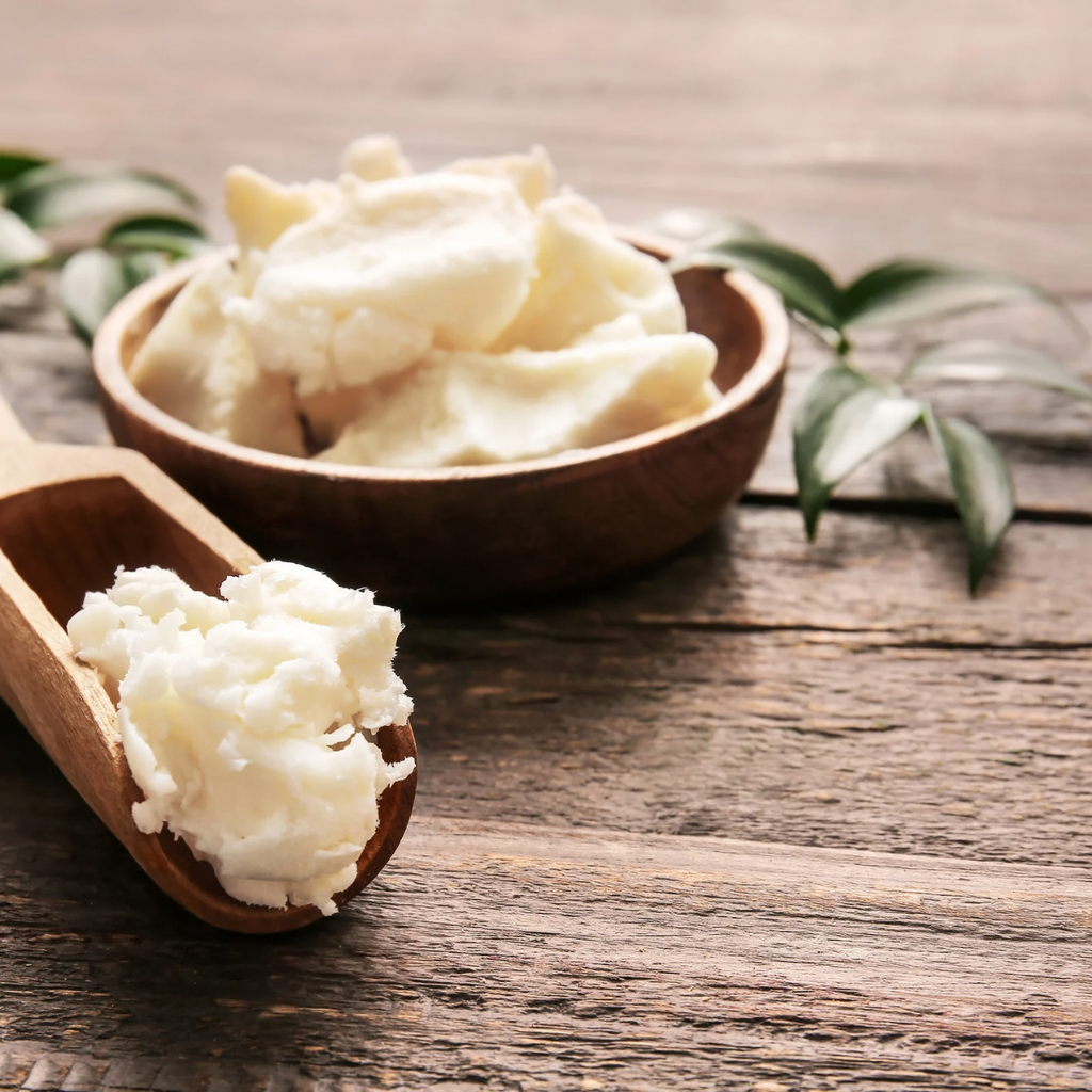 Raw shea butter on a wooden scoop and in a rustic wooden bowl, displayed on a wooden table with fresh green leaves — natural skincare ingredients.